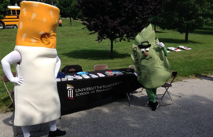 Anti-smoking mascots stand in front of the table promoting the School of Pharmacy at the Movin' and Groovin' Health Fair