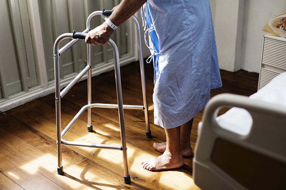 Elderly patient stands near hospital bed grasping walker.